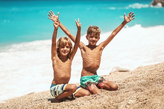 Little brothers with raised arms are having fun at the tropical beach and looking at camera.