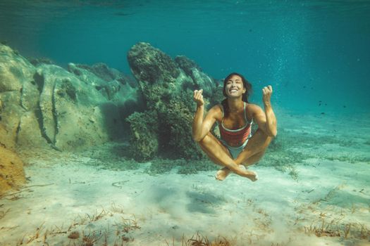 A beautiful young woman is having fun at summer vacation exploring seafloor during scuba diving in sea. She is doing yoga under the sea.
