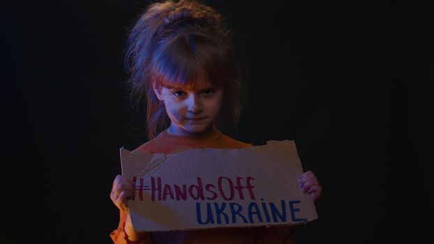 Upset poor toddler dirty faced homeless girl kid protesting war conflict raises banner with inscription massage text on cardboard Hands Off Ukraine on black background. Peace, no war, stop aggression