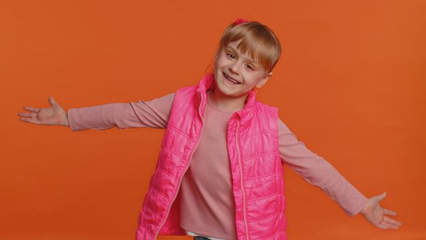 Funny joyful sincere little girl 6 years old in vest making playful silly facial expressions and grimacing, fooling around showing tongue. Young toddler child isolated alone on orange wall background