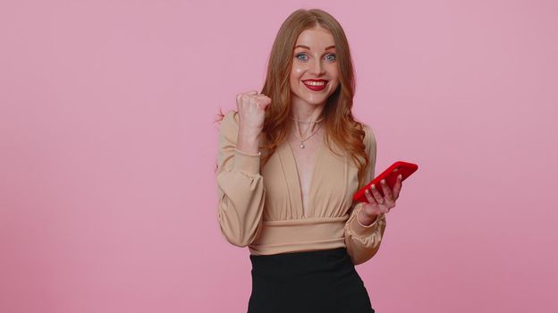 Happy excited joyful businesswoman girl in blouse use mobile cell phone typing browsing say wow yes found out great big win news doing winner gesture. Young adult woman on pink studio wall background