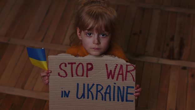 Scared Ukrainian homeless toddler girl sitting on floor at home holding inscription massage Stop War In Ukraine. Child alone hiding from bombing attack. Crisis, peace, no war, Russian aggression