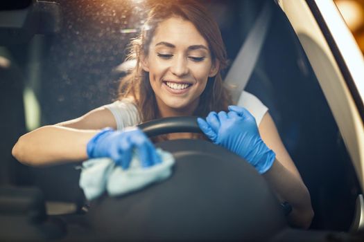 A young smiling woman with protective gloves on her hands, wipes a steering wheel in with a cloth ready to hit the road. 