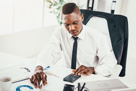 Pensive African businessman working on calculator in modern office and planning what to do next.
