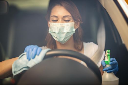 A young woman with a mask on her face  and protective gloves on her nands wipes a steering wheel in her car holding a cloth in one and a bottle with disinfectant in the other hand.
