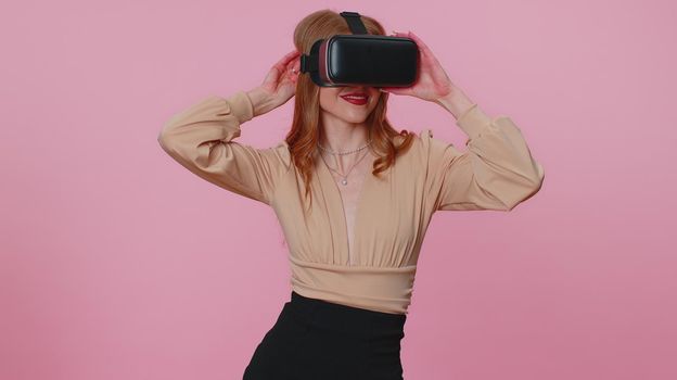 Amazed teen student girl using headset helmet app to play simulation game. Watching virtual reality 3D 360 video. Young woman in VR goggles isolated on pink background. Addiction from technology