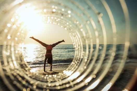 A beautiful creative composition of a sea landscape shot through a circle focus showing a teenage girl who makes gymnastic figure on the beach in sunset.