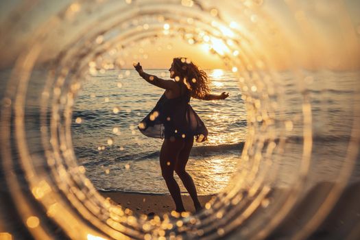 A beautiful creative composition of a sea landscape shot through a circle focus showing a young woman who is dancing on the beach in sunset.