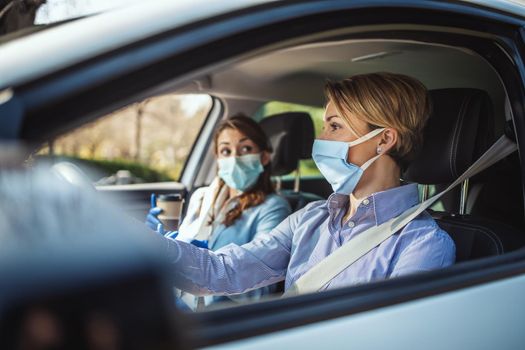 Young business women with protective masks on their faces are going to the work together by car and talking about global crisis. 
