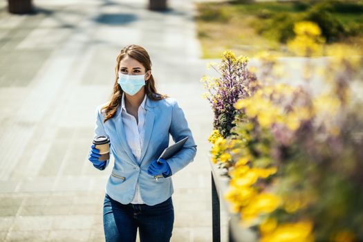 Young business woman with protective mask on her face and digital tablet and coffee in her hands is going to work at covid 19 pandemic.