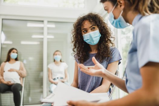 Upset African American teenage girl with protective mask talks with nurse while waits for gynecologist check up.