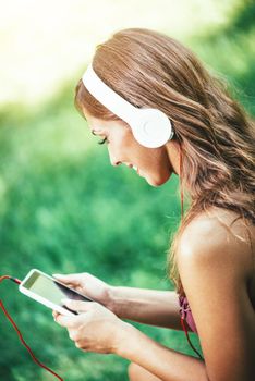 Woman relaxes with headphones listening to music sitting in park. She is texting messages on her smartphone