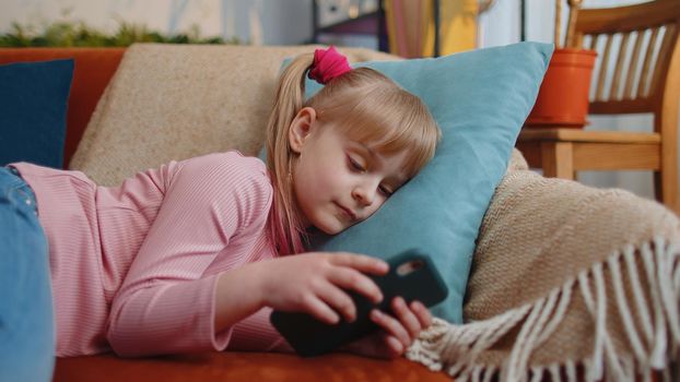 Child toddler hold smartphone watching funny cartoons, chatting with friends lying on couch alone at home. Little one school kid girl using mobile phone technology device. Children tech addiction