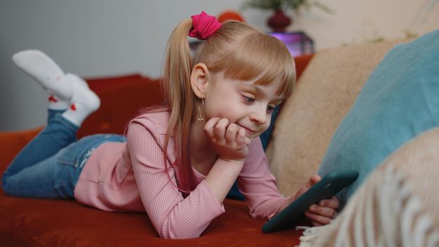 Little one school kid girl children using mobile phone technology device lying on couch alone at home. Child toddler hold smartphone watching funny cartoons, chatting, play online game. Tech addiction