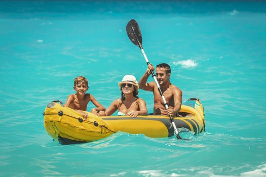 Happy family is enjoying a ride in rubber kayak and waves are splashing them at tropical ocean water during summer vacation.
