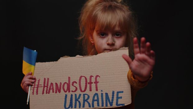 Scared Ukrainian poor dirty toddler girl homeless protesting war conflict raises banner inscription cardboard massage Hands Off Ukraine. Crisis, no peace, stop Russian aggression. Child against war