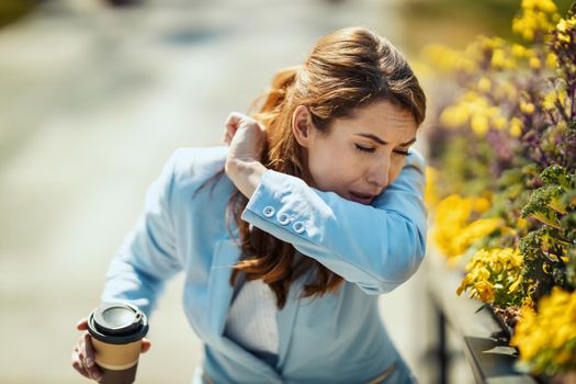 A young businesswoman on a coffee break outside is sneezing into the elbow by an allergy or a cold.