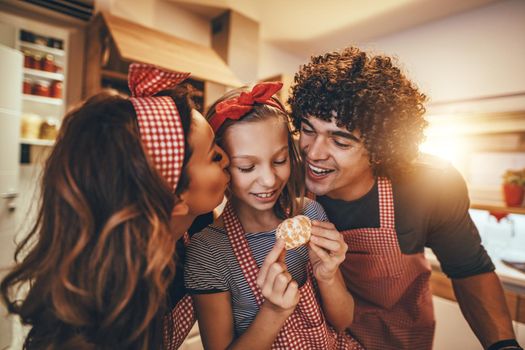 Happy parents and their daughter enjoy and having fun in making and having healthy meal together at their home kitchen. Parents are kissing a little girl while she is holding a mandarin ready to eat.