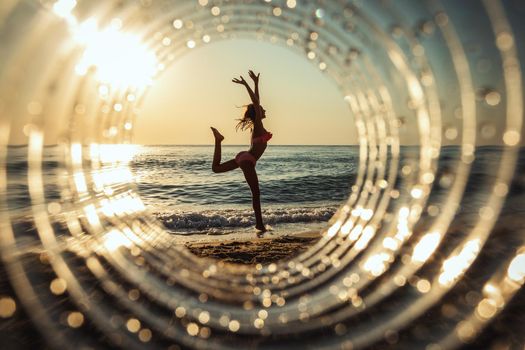 A beautiful creative composition of a sea landscape shot through a circle focus showing a teenage girl who makes gymnastic figure on the beach in sunset.