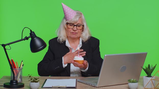 Sad upset lonely senior businesswoman in festive hat celebrating birthday party alone, feel frustrated despair disappointed sadness holds cake with candle at office. Elderly woman on green chroma key