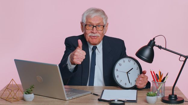 It is your time. Senior businessman showing time on wall office clock, ok, thumb up, approve, pointing finger at camera. Elderly man sits at office desk isolated indoor on pink studio wall background