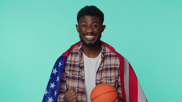 African american one young man 20s sportsman basketball fan in checkered shirt holding American USA flag doing winner gesture, dancing isolated on blue background. Workout sport motivation lifestyle