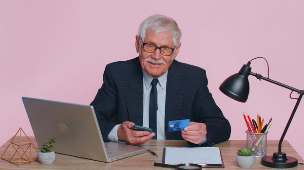 Senior businessman using credit bank card and smartphone while transferring money, purchases online shopping sits at office workplace desk. Elderly man isolated indoor on pink studio wall background