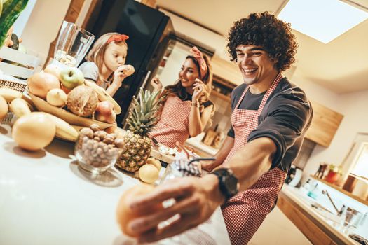 Happy parents and their daughter cooking together in the kitchen while little girl eating mandarin.