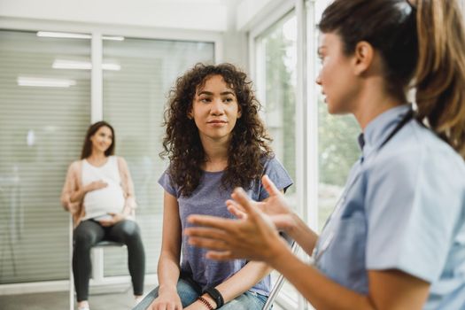 Upset African American teenage girl talks with nurse while waits for gynecologist check up.