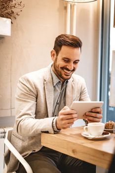 Handsome young smiling man drinking coffee in a cafe. He is surfing the internet on a digital tablet.