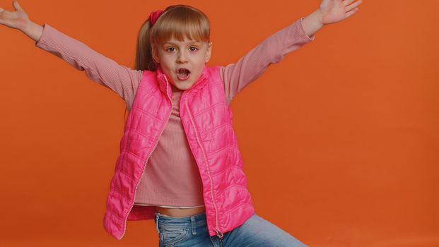 Funny happy sincere little girl 6 years old in vest making playful silly facial expressions and grimacing, fooling around showing tongue. Young toddler child isolated alone on orange wall background