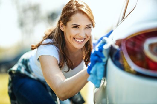 A smiling young woman with protective gloves on her nands wipes her car with a cloth ready to hit the road.