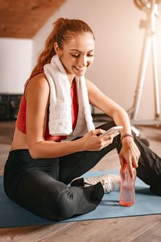 Attractive woman using a smartphone while taking break of exercising in living room at home.