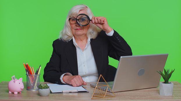 Investigator researcher scientist senior businesswoman hold magnifying glass near face, look into camera with big zoomed funny eye, search, analysing sits at workplace desk at green chroma key office