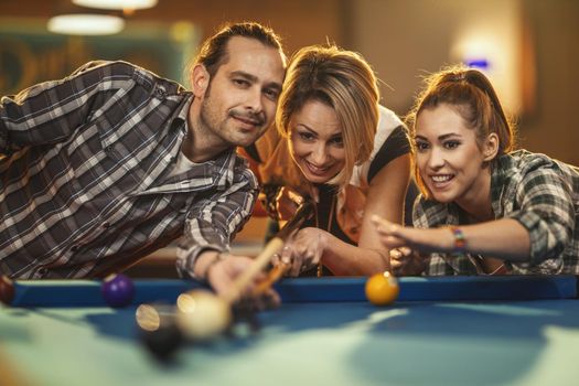 Group of a smiling cheerful friends are playing billiards in bar after work. They are involved in recreational activity.