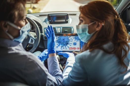 Young business women with protective masks on their faces are sitting in a car and talking about global crisis. They are showing each other an animation of a corona virus on a tablet.