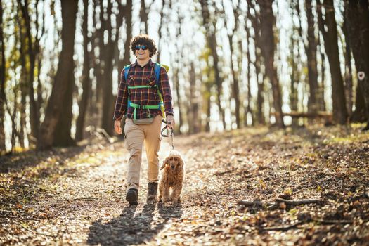 Happy smiling handsome man is walking along the forest path with his dear cute dog enjoying landscape in forest in beautiful sunny day.