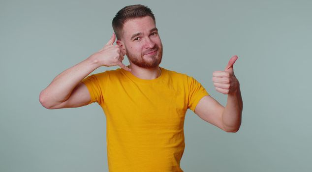 Call me, here is contact number. Cheerful one man in yellow t-shirt looking at camera doing phone gesture like says hey you call me back. Young guy posing isolated alone on gray studio wall background