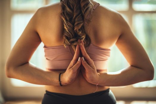 Close-up of a young woman is doing yoga Back Namaste pose in the living room at home. She is meditating in morning sunshine.