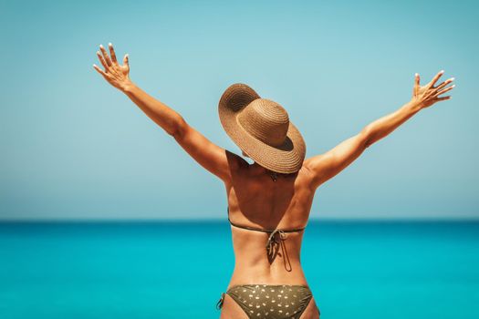Handsome midle age woman in bikini wearing straw hat and enjoying sunbathing at beach by the ocean with open arms.