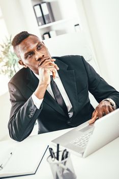 Pensive African businessman working on laptop in modern office.