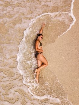 Aerial view picture from drone of woman relaxing on the idyllic tropical beach. She is laying on the sandy beach while the waves splash on it. 