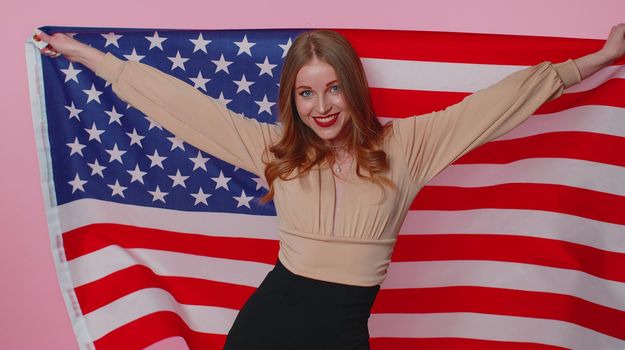 Trendy cheerful positive teen girl waving and wrapping in American USA flag, celebrating, human rights and freedoms. Independence day. Young pretty adult woman. Indoors studio shot on pink background