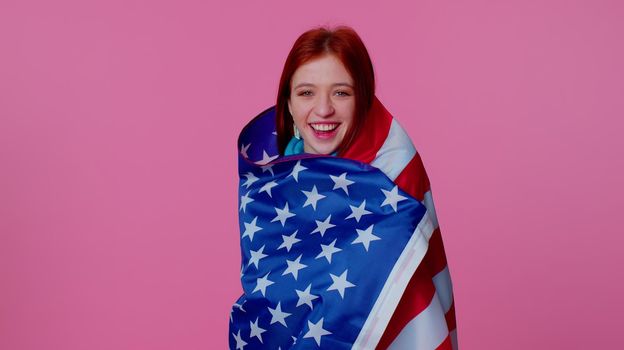 Trendy cheerful positive teen girl waving and wrapping in American USA flag, celebrating, human rights and freedoms. Independence day. Young pretty adult woman. Indoor studio shot on pink background