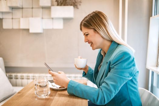 Young smiling businesswoman on a break in a cafe. He is drinking coffee and using smartphone. 