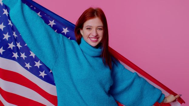 Trendy cheerful positive teen girl waving and wrapping in American USA flag, celebrating, human rights and freedoms. Independence day. Young pretty adult woman. Indoor studio shot on pink background