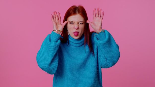 Cheerful funny portrait of teenager student girl in blue sweater showing tongue making faces at camera, fooling around, joking, aping with silly face, teasing. Young woman on pink studio background