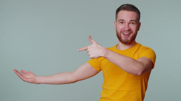Handsome one bearded man showing thumbs up and pointing empty place, advertising area for commercial text, copy space for goods promotion. Young adult guy. Studio shot indoors on gray wall background