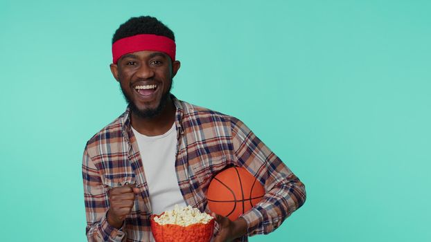 Bearded happy young african american man 20s sportsman alone basketball fan in shirt holding ball doing winner gesture, dancing isolated on blue background. Workout sport motivation lifestyle concept