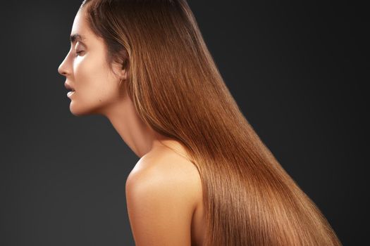 Beautiful yong woman with long straight brown hair on dark background. Sexy fashion model with smooth gloss hairstyle, keratin treatment